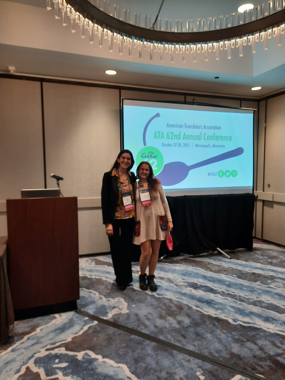 La fotografía muestra a Maitén Vargas y Erika Cosenza de pie al lado de un atril en una sala de conferencias vacía. Detrás de ellas, una pantalla muestra el logo de la ATA 62nd Annual Conference.