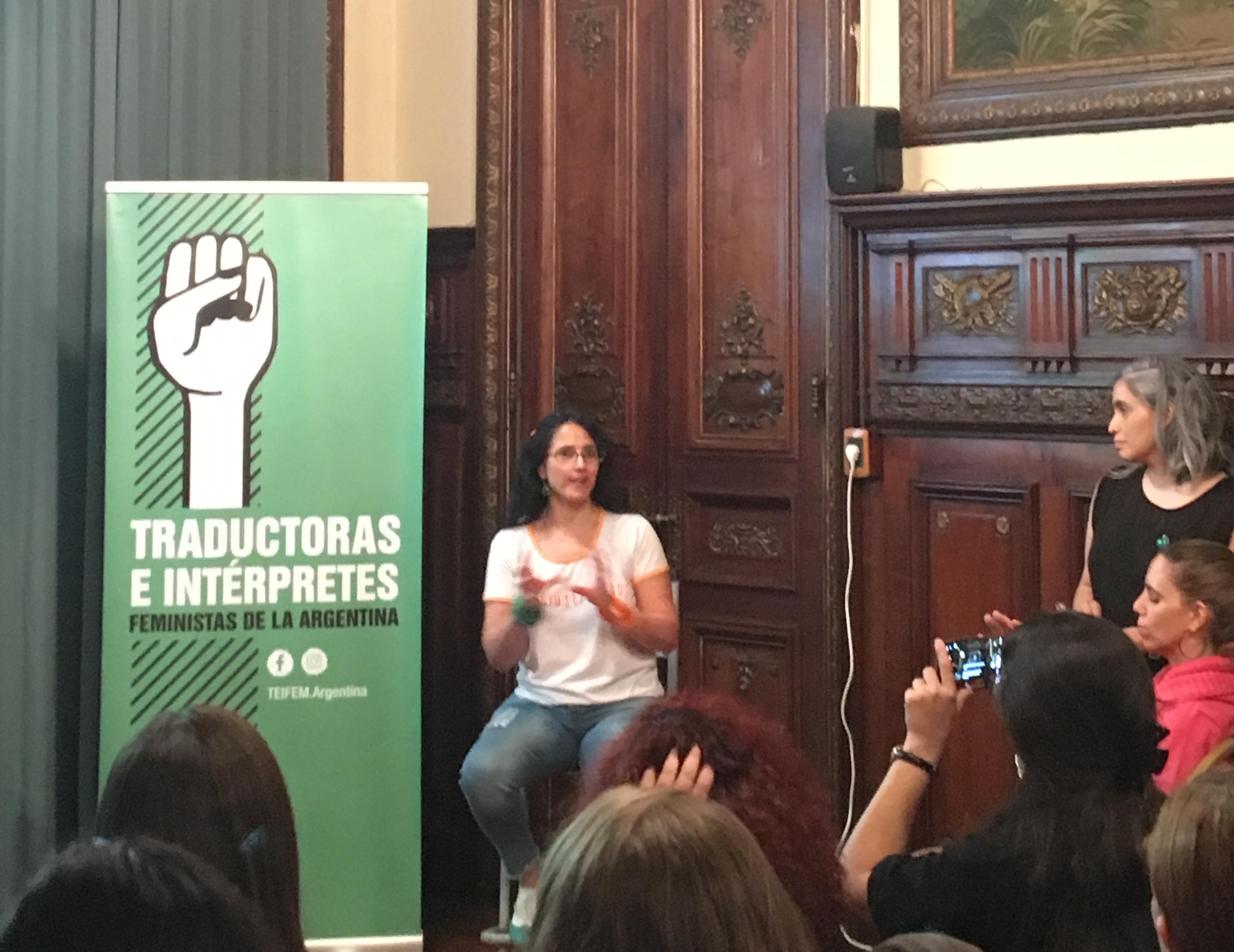 La fotografía muestra a Erika Cosenza sentada en un taburete alto al lado de un banner vertical que, sobre fondo verde, dice «Traductoras e Intérpretes Feministas de la Argentina»