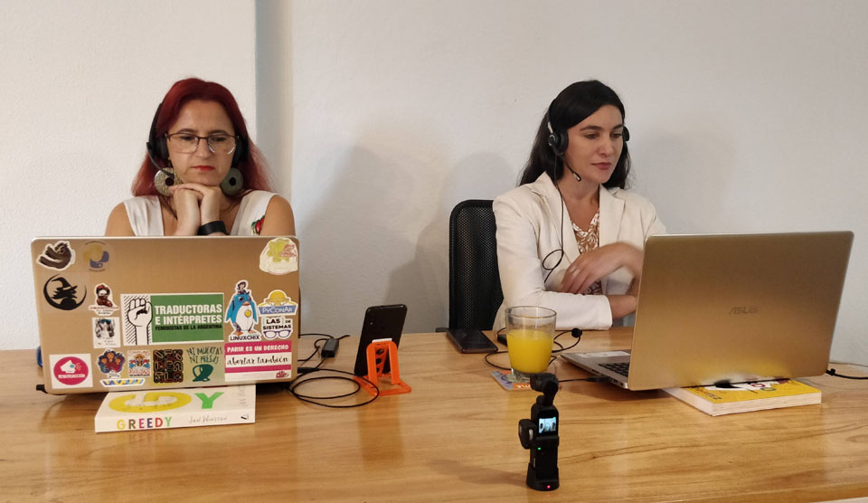 La fotografía muestra a Maitén Vargas y Erika Cosenza sentadas frente a sus respectivas laptops. Hay una cámara que las está filmando.
